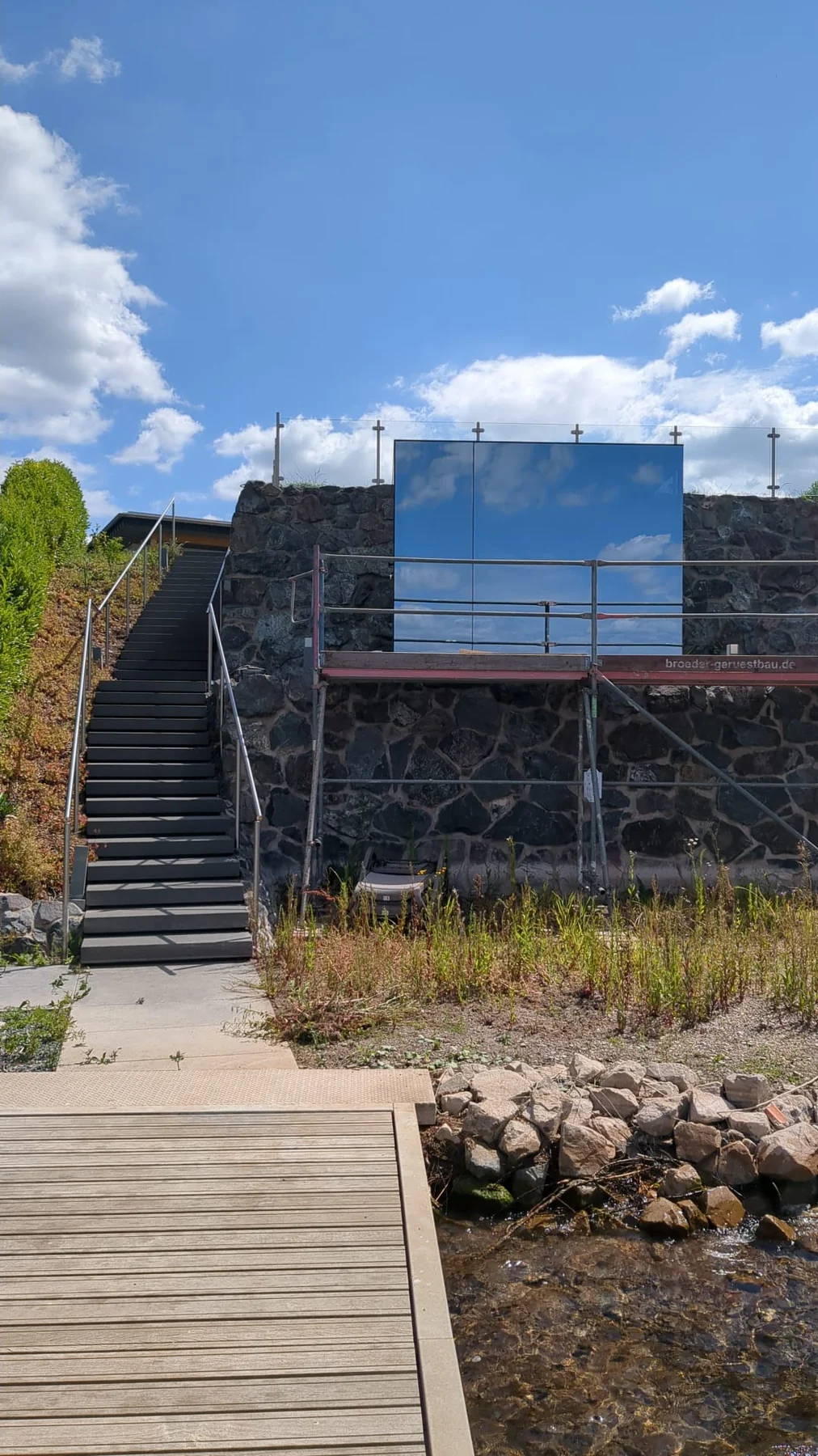 Mirror+ exterior during installation against a stone wall, reflecting blue sky and clouds