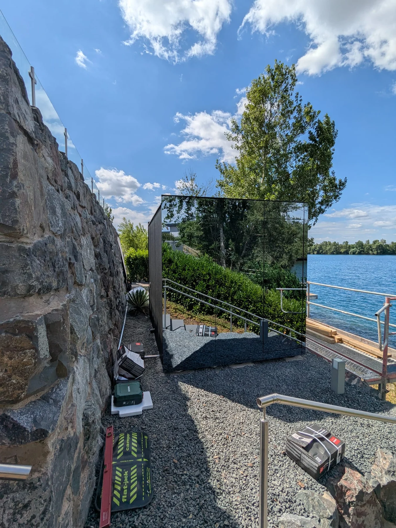 Mirror+ exterior during installation beside a natural stone wall and waterfront, reflecting nearby greenery
